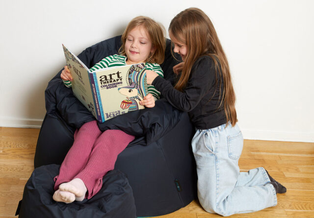 young girl reading a book on the protac sensit jr chair with another young girl next to her smiling